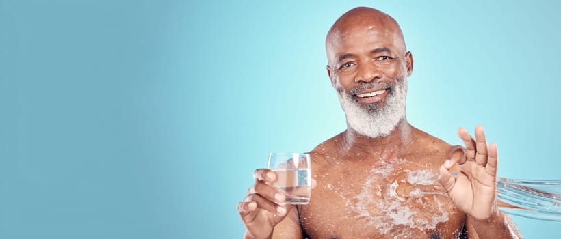 Water splash, portrait and senior man in a studio with a glass of aqua and a ok hand gesture. Health, wellness and happy elderly African male with sign language by a blue background with mockup space.