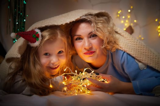 Cute mother and daughter in pajamas lie on soft blanket and having fun in room with Christmas garlands and white background. Tradition of decorating house for holiday. Happy childhood and motherhood