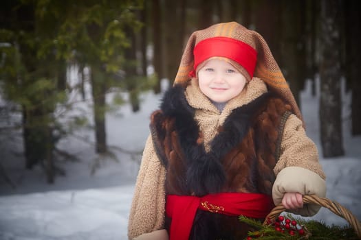Little girl in thick coat and a red sash with basket of fir branches and red berries in cold winter day in the forest. Medieval peasant girl with firewood. Photoshoot in stile of Christmas fairy tale