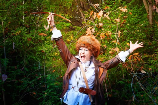 Girl in a leather jacket, a big red fox fur hat and with a crossbow in the forest in autumn. A female model poses as a fabulous royal huntress on nature hunt at a photo shoot