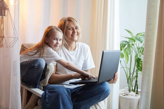 Beautiful young woman and her little cute daughter are using laptop at home. Enjoying spending time together with Internet and modern technologies