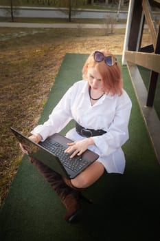 Beautiful redhead moden girl working by laptop in gazebo on autumn, summer or spring day. Businesswoman, student, freelancer or manager works outdoors in natural landscape in a village, city or town