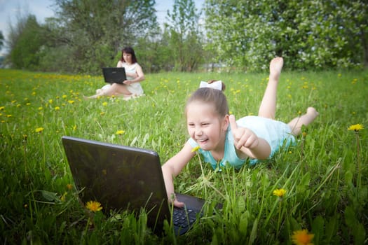 Young girl with laptop resting on a green lawn or a field in a park in nature and her mon on background. Fmily with mother and daughter chatting, working, communicating in Internet