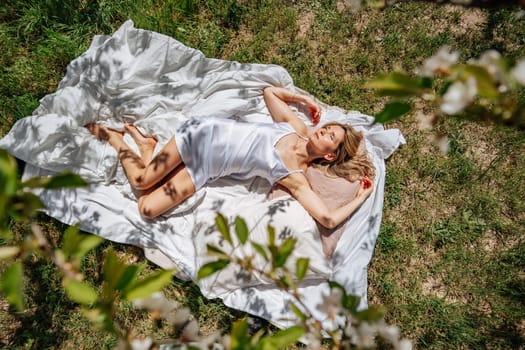 woman sleeps on a white bed in the fresh spring grass in the garden. Dressed in a blue nightgown