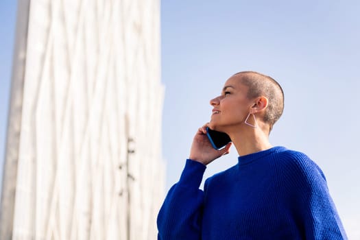 confident woman with buzz cut in blue sweater talking happy on mobile phone at city, concept or modern lifestyle and technology of communication, copy space for text