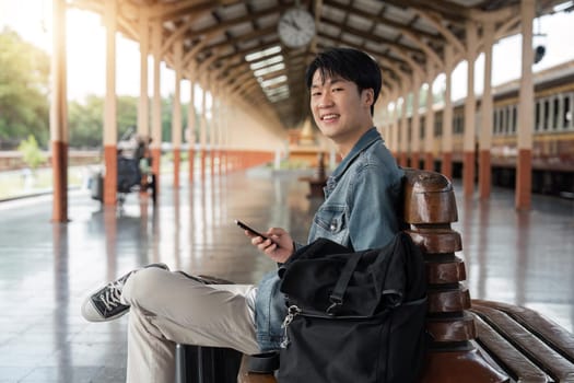 Traveler young asian man using cellphone booking trip at train station. Happy tourist travel by train using smartphone searching location. Male Backpacker arrival at platform railway.