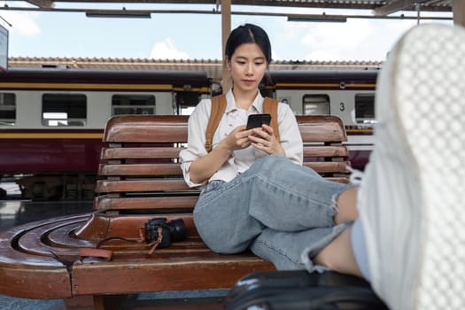 Traveler young asian woman using cellphone trip at terminal train station. Happy tourist travel by train using smartphone searching location. Female Backpacker arrival at platform railway.
