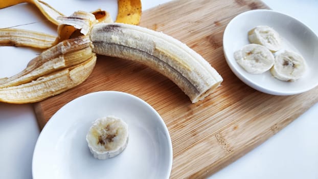 Banana with open panel on wooden board and sliced round pieces on the plate on white background. Ripe banana with peel and cooking. Delicious sweet fruit dessert. Close up