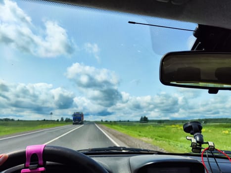 view through car windshield of natural landscape with green field, white clouds in blue sky. Hand of woman on steering wheel of car. Female traveler driving on trip or journey in spring or summer