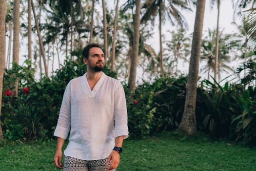 Close up shot of young bearded man in white shirt on green palms background. Male tourist in tropical jungle