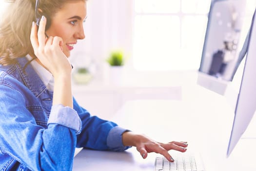 Happy charming young woman sitting and working with laptop using headset in office.