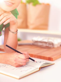 Happy beautiful woman standing in her kitchen writing on a notebook at home.