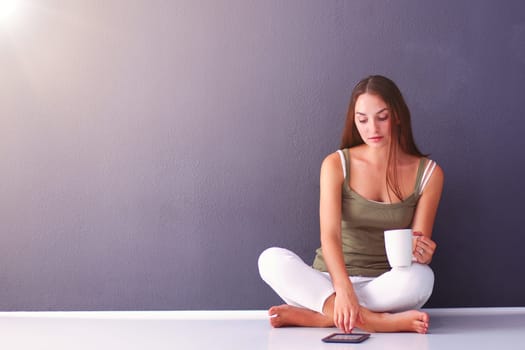 Attractive caucasian girl sitting on floor with cup and tablet near wall.