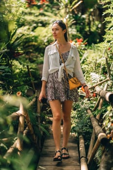 Front view of walking girl on wood bridge in jungle. Beautiful lady is summer stylish outfit exploring beauty of balinese nature