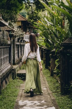 Back view of walking girl in the garden temple. Tropical vegetation and balinese architecture wall