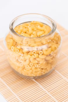 Uncooked Chifferi Rigati Pasta in Glass Jar on Bamboo Mat on White Background. Fat and Unhealthy Food. Classic Dry Macaroni. Italian Culture and Cuisine. Raw Pasta