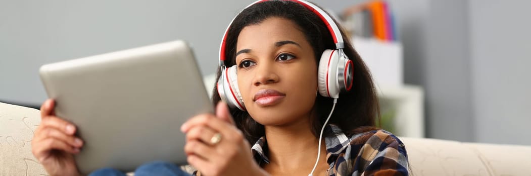 Focused black woman studying foreign language online. Remote female student in virtual classroom sitting on sofa in headset and holding tablet