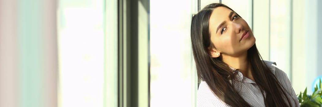 Dreamy young business woman is sitting on windowsill near huge window in modern office sun is shining brightly. Businesswoman enjoying break and relaxing after a hard day at work