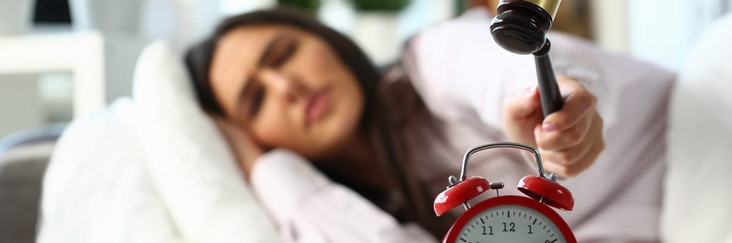 Young woman tries to smash an alarm clock with wooden mallet and destroy clock. Displeased angry girl lies in bed and turns off the alarm clock with hammer