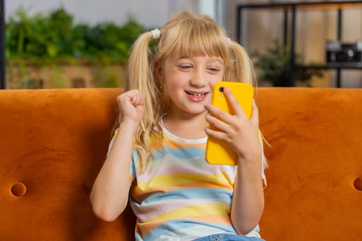 Young blonde child kid girl use mobile smartphone typing playing browsing say Wow yes found out great big surprise, good news, celebrate online game win. Female toddler at home in room sits on sofa