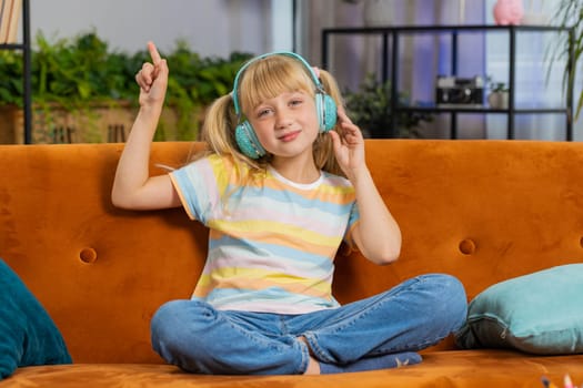 Happy overjoyed toddler girl kid in wireless headphones relaxing sitting on couch at home room choosing listening favorite energetic disco dancing music. Children weekend leisure activities. Lifestyle