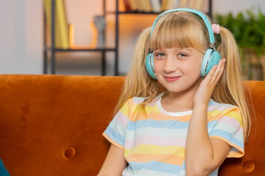 Happy overjoyed toddler girl kid in wireless headphones relaxing sitting on couch at home room choosing listening favorite energetic disco dancing music. Child weekends leisure activities. Lifestyles