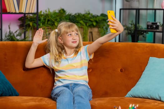 Young blonde child kid girl blogger taking selfie on smartphone, communicating video call online with social media subscribers. Female teen toddler recording video story at home in room sits on sofa