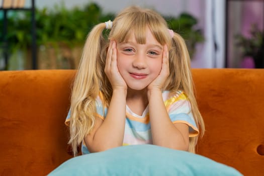 Close-up portrait of happy smiling Caucasian preteen school girl 7 years old. Young blonde lovely child kid looking at camera at home play room apartment sitting on orange couch. Ponytails hairstyle