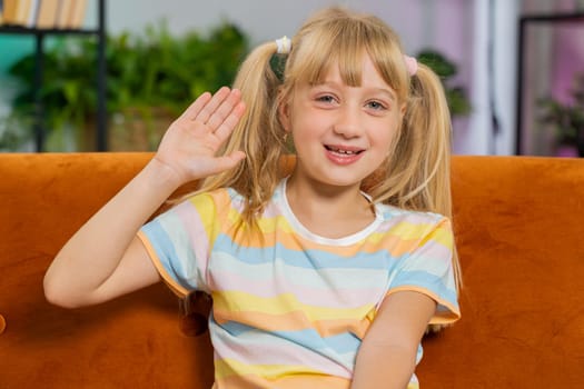 Young blonde child kid girl smiling friendly at camera and waving hands gesturing hello, hi, greeting or goodbye, welcoming with hospitable expression. Teen toddler at home in living room sits on sofa