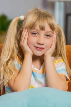 Close-up portrait of happy smiling Caucasian preteen school girl 7 years old. Young blonde lovely child kid looking at camera at home play room apartment sits on couch. Ponytails hairstyle. Vertical