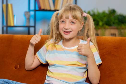 Like. Happy young blonde child kid girl looking approvingly at camera showing thumbs up, like sign positive something good, positive feedback. Female teen toddler at home in living room sits on sofa