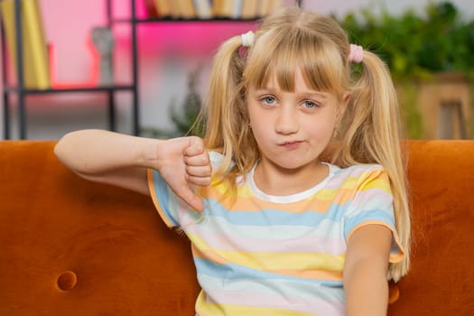Dislike. Upset girl showing thumbs down sign gesture, expressing discontent, disapproval, dissatisfied bad work at modern home apartment indoors. Displeased teen child, kid in living room on sofa