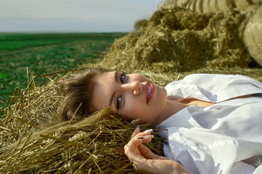 A young brunette woman in a white shirt and jeans lies on hay rolls. Nature, summer holidays, vacation concept - young woman lies and dreams in the grain field