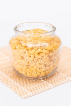 Uncooked Chifferi Rigati Pasta in Glass Jar on Bamboo Mat on White Background. Fat and Unhealthy Food. Classic Dry Macaroni. Italian Culture and Cuisine. Raw Pasta