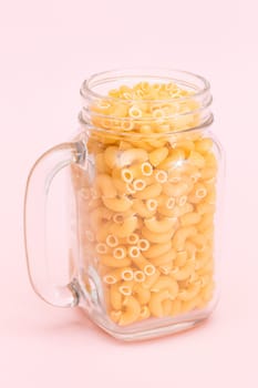 Uncooked Chifferi Rigati Pasta in Glass Jar on Pink Background. Fat and Unhealthy Food. Scattered Classic Dry Macaroni. Italian Culture and Cuisine. Raw Pasta