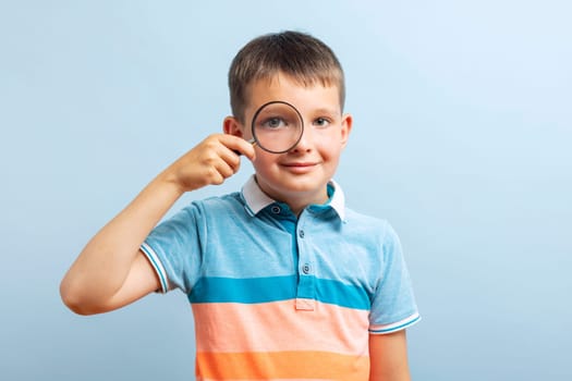 Positive curious schoolboy in casual clothes looks through magnifying glass on a blue backgrounds.