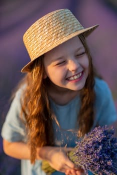 Lavender sunset girl. A laughing girl in a blue dress with flowing hair in a hat walks through a lilac field, holds a bouquet of lavender in her hands