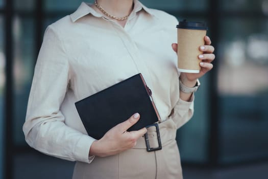 Close-up on a notebook for notes and a paper cup with coffee in the female hands of an office worker.