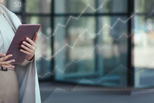 Digital tablet in female hands close-up against the background of an office building and charts growing up. Successful startup concept.