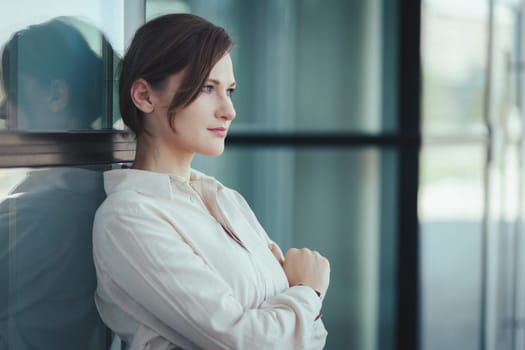 A self-confident beautiful young caucasian woman stands at the office building leaning against a glass wall, purposefully thinking looks to the side.