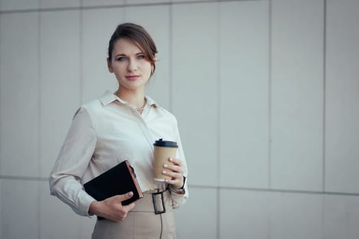 Caucasian young woman in office or business fashion style in clothes with notebook and paper cup with look at camera, copy space.