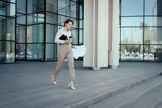 Purposeful business woman with coffee notepad and documents hurry coming out of the office building.