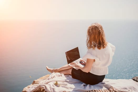 Successful business woman in yellow hat working on laptop by the sea. Pretty lady typing on computer at summer day outdoors. Freelance, travel and holidays concept.