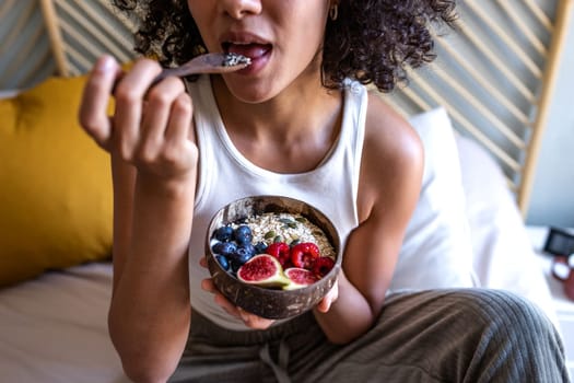 Unrecognizable young multiracial woman eating healthy breakfast bowl of oats and fruit. Breakfast in bed. Healthy lifestyle concept.