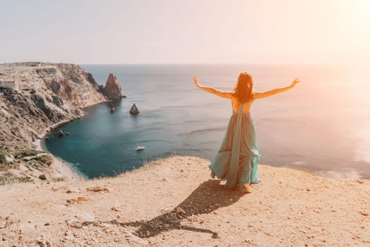 Side view a Young beautiful sensual woman in a red long dress posing on a rock high above the sea during sunrise. Girl on the nature on blue sky background. Fashion photo.