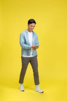Happy man in casual wear using mobile phone chatting online, walking on yellow background