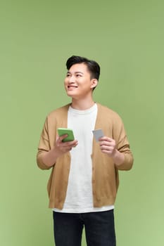 Smiling man holding credit card and mobile phone while standing against green background