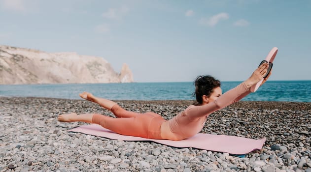 Middle aged well looking woman with black hair doing Pilates with the ring on the yoga mat near the sea on the pebble beach. Female fitness yoga concept. Healthy lifestyle, harmony and meditation.