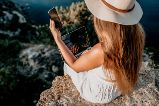 Successful business woman in yellow hat working on laptop by the sea. Pretty lady typing on computer at summer day outdoors. Freelance, travel and holidays concept.