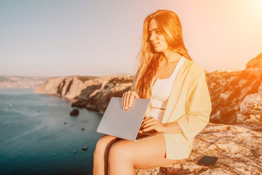 Successful business woman in yellow hat working on laptop by the sea. Pretty lady typing on computer at summer day outdoors. Freelance, travel and holidays concept.